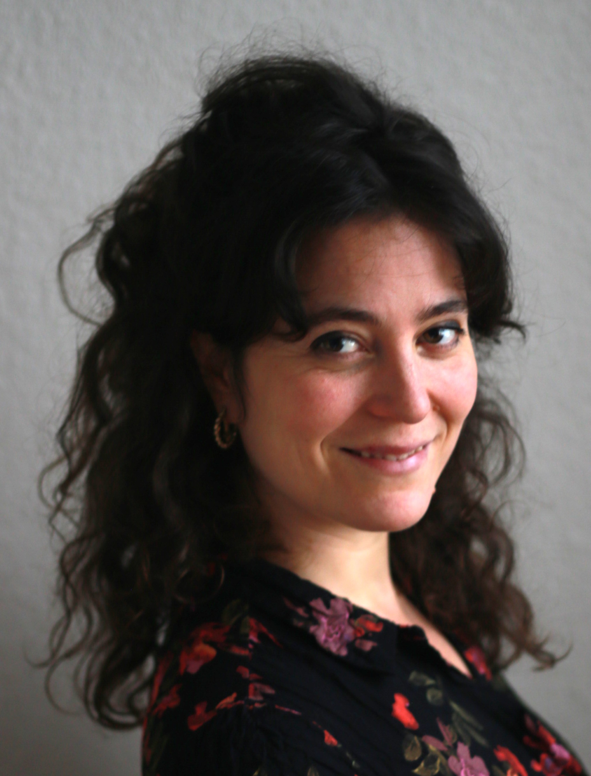 Weiße Frau vor grauer Wand, schaut über rechte Schulter zur Kamera. Hat schwarze Locken, die halb zu einem Zopf gebunden sind. Trägt schwarze Bluse mit roten Blumen.