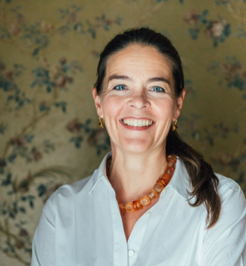 Weiße Frau mit braunen Haaren, trägt ein weißes Hemd und eine große Bernsteinkette. Vor Blümchentapete.