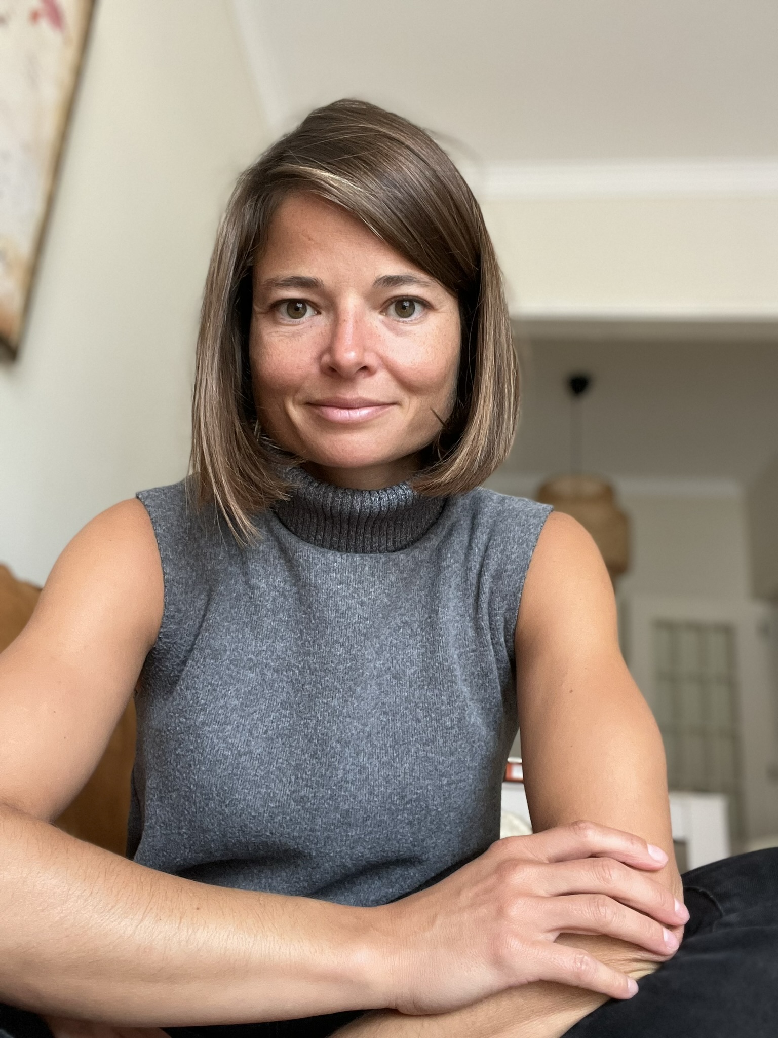 Weiße Frau mit dunkelblondem Bob. Trägt ein ärmelloses graues Rollkragen-Oberteil. Arme sind verschränkt auf Beinen abgestützt.